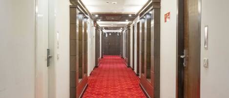 Um corredor de hotel com carpete estampado em vermelho, portas em ambos os lados e teto com iluminação embutida.