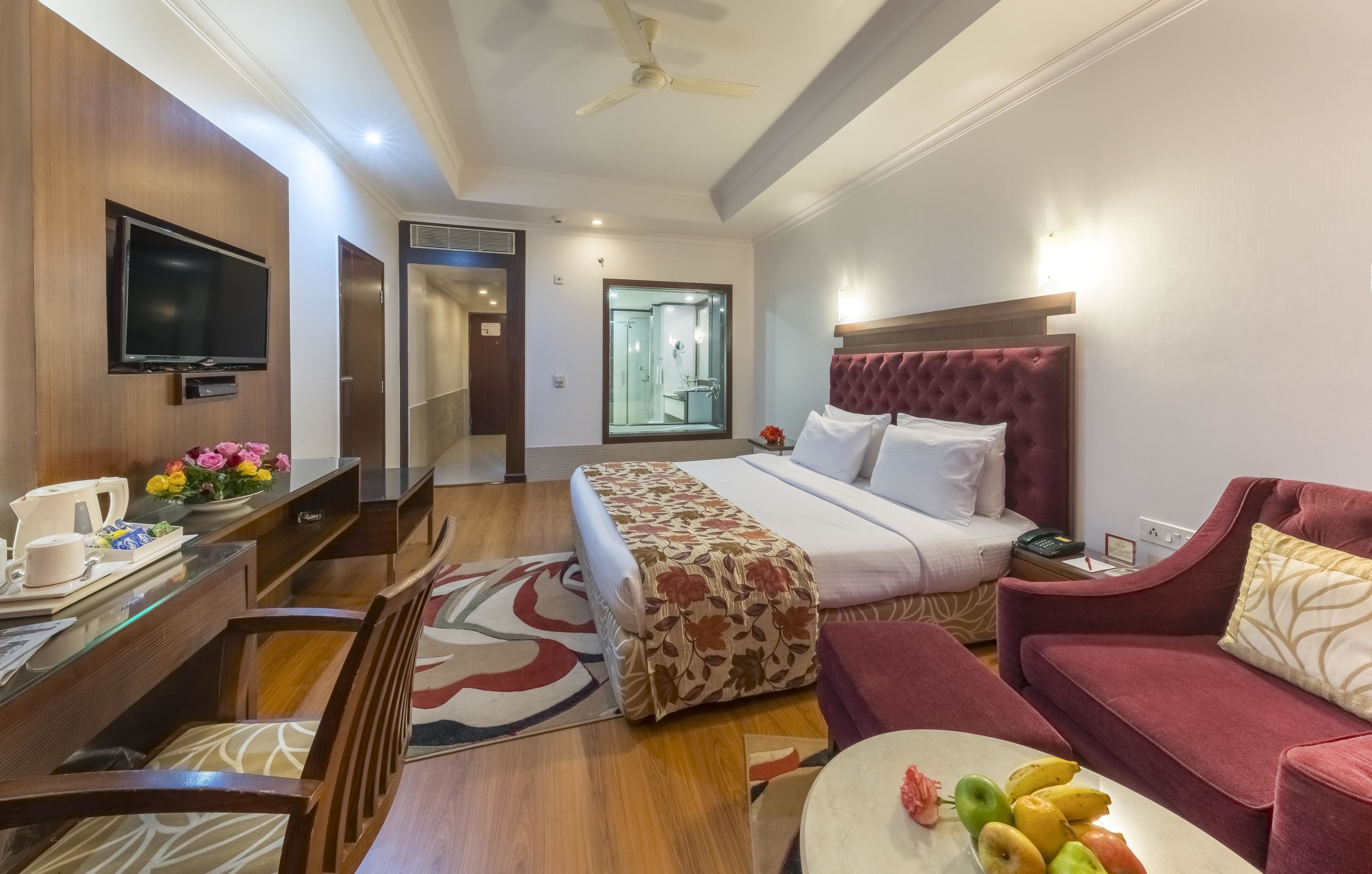 A hotel room with a bed, a desk with a TV, a red sofa, and a dining table with fruit.