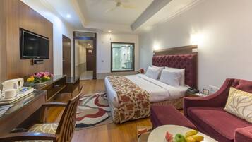 A hotel room with a bed, a desk with a TV, a red sofa, and a dining table with fruit.