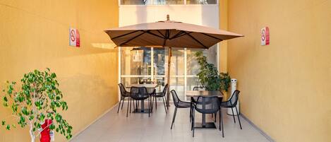 Un patio exterior con mesas y sillas, una sombrilla grande y una planta en maceta.