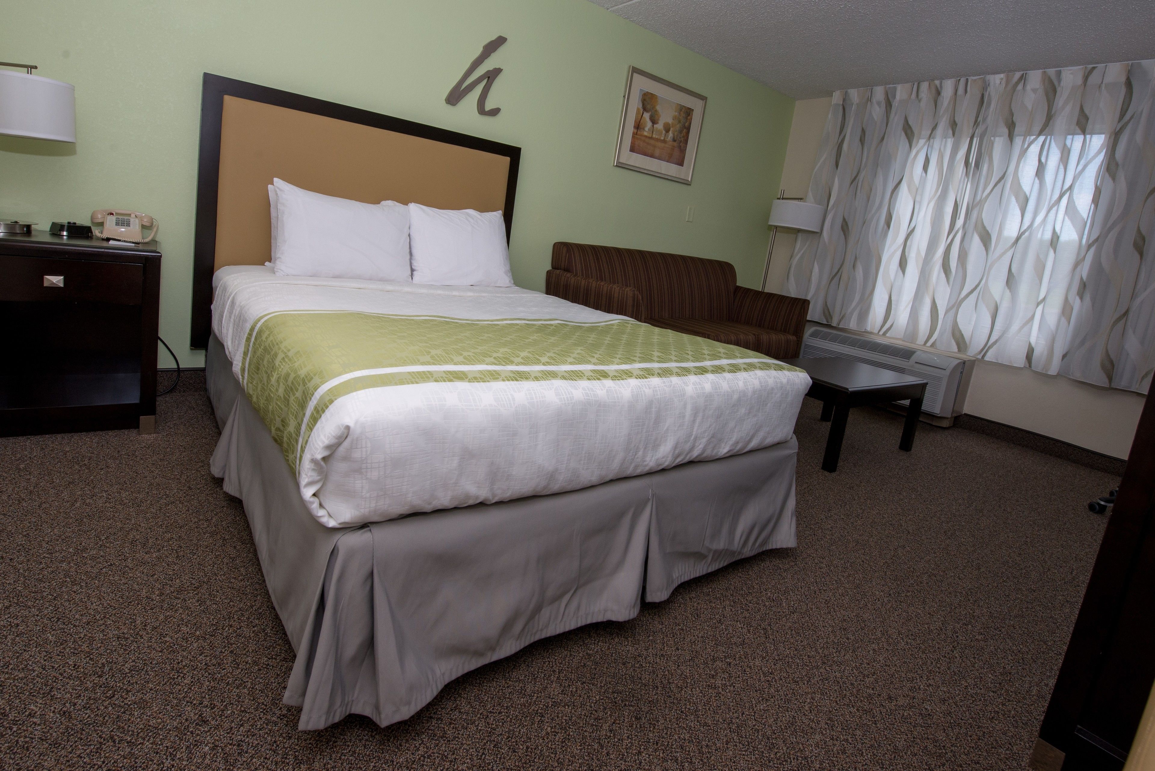 A hotel room with a large bed, a bedside table, a chair, a desk, a lamp, and a picture on the wall.