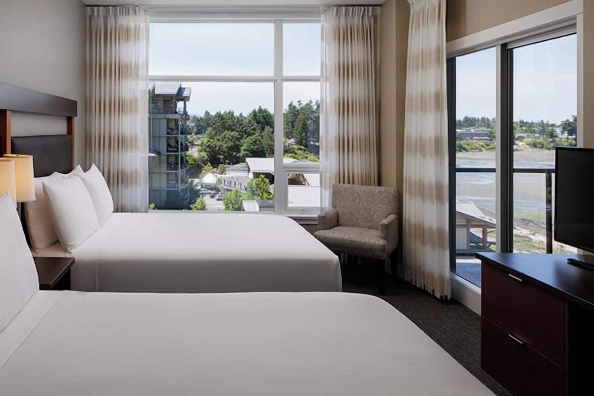 A hotel room with two beds, a chair, a TV, and a large window offering a view of buildings and greenery.