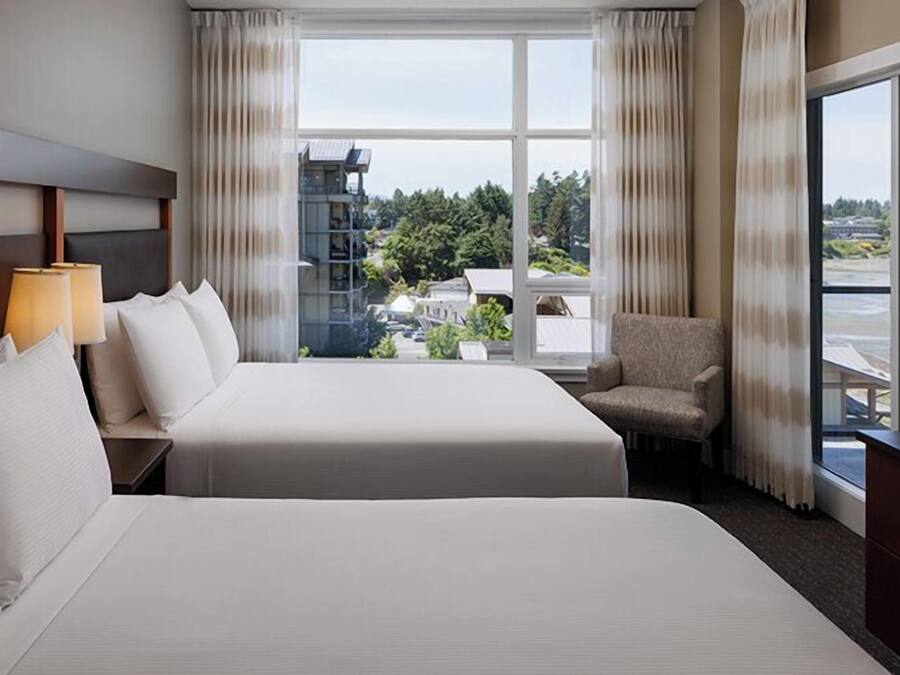 A hotel room with two beds, a chair, a TV, and a large window offering a view of buildings and greenery.