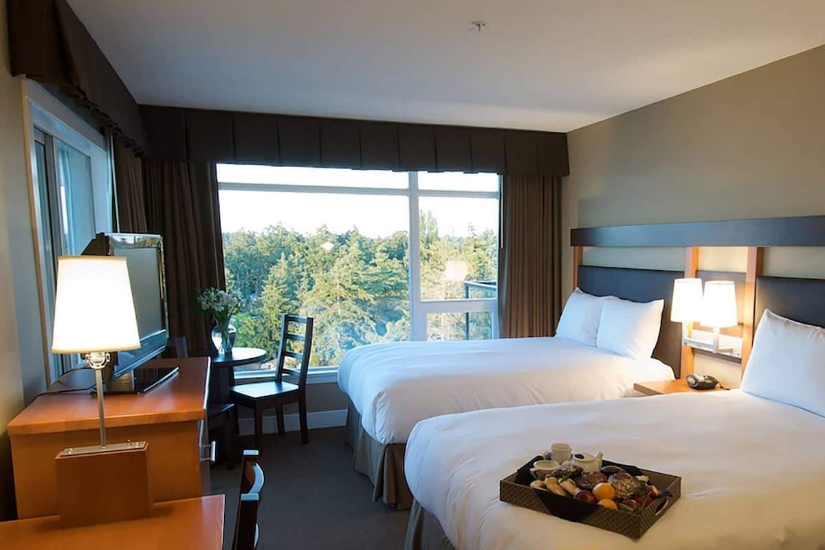 A hotel room with two beds, a desk with a lamp, a chair, and a window with a view of trees.
