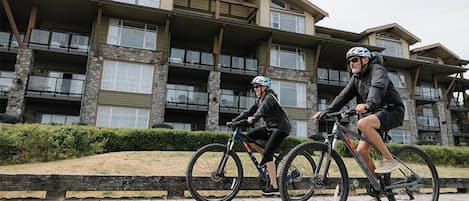 Two people riding bicycles on a wooden deck in front of a multi-story building with balconies and stone accents.