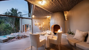 Un área de comedor al aire libre con una mesa dispuesta para dos personas, rodeada de sillas con cojines, una terraza de madera y un muro de piedra.