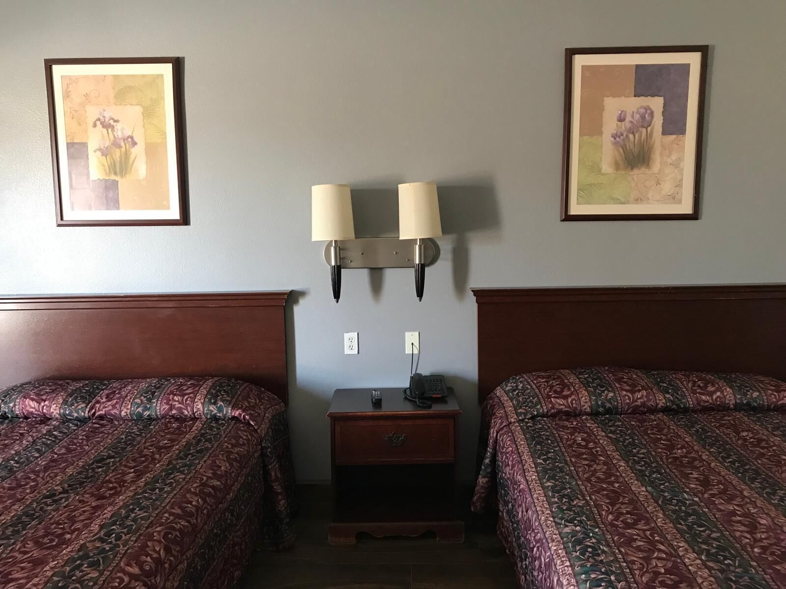Habitación de hotel con dos camas, cada una con una colcha estampada, una mesita de noche con lámpara y dos cuadros florales enmarcados en la pared.
