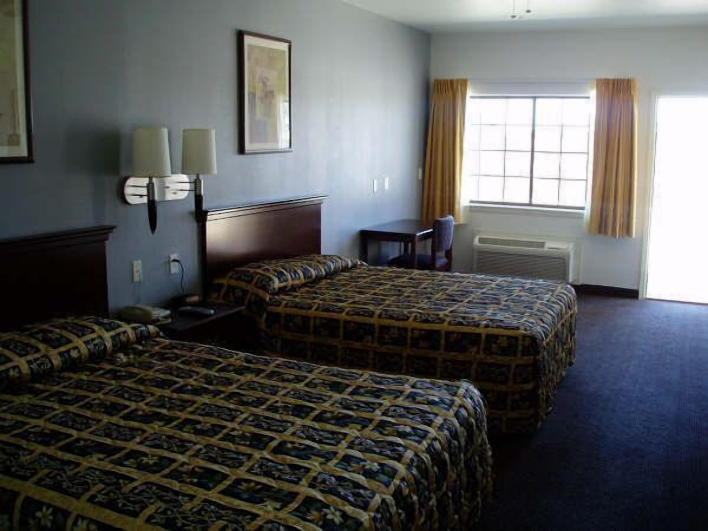 A hotel room with two beds, a desk, a chair, a window with curtains, and a framed picture on the wall.
