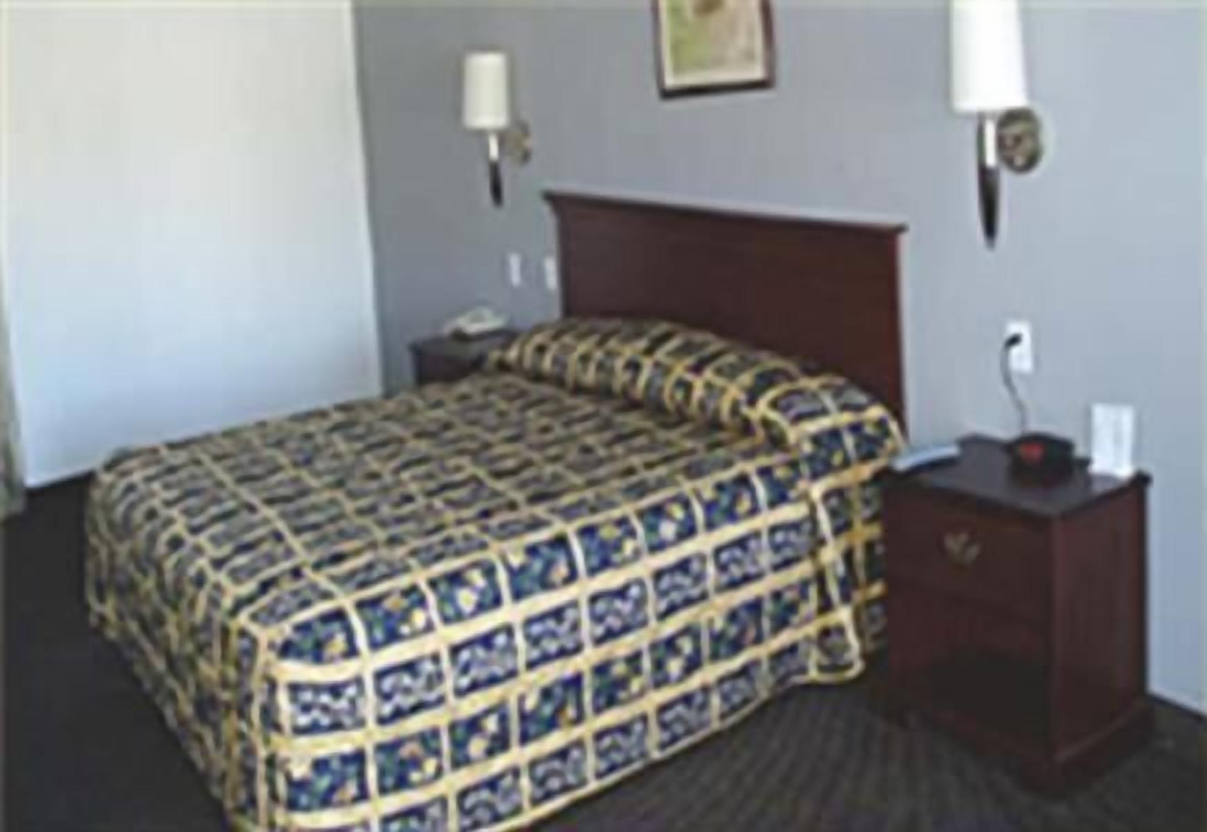 Quarto de hotel com cama, mesa de cabeceira, luminária e relógio fixado na parede.