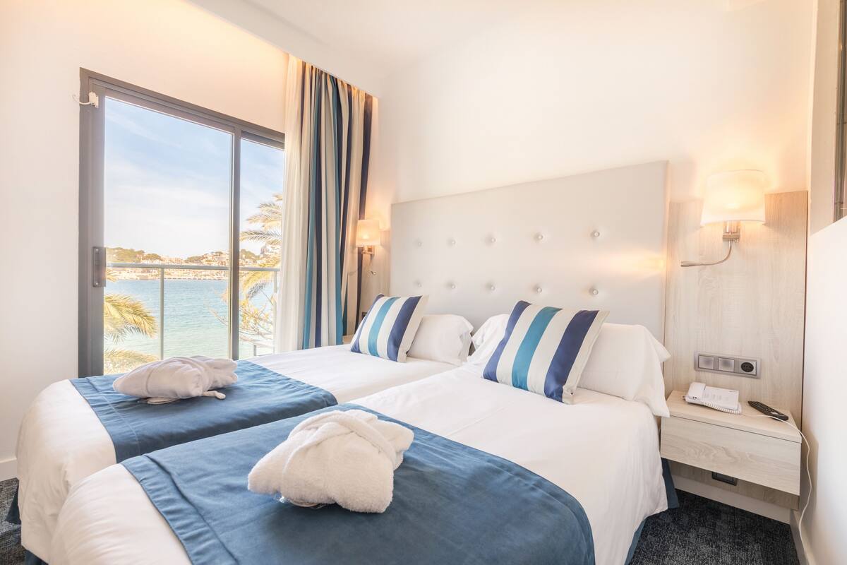 A hotel room with two beds, a large window with a view of the sea, a bedside table with a lamp, and a nightstand with a phone and remote control.