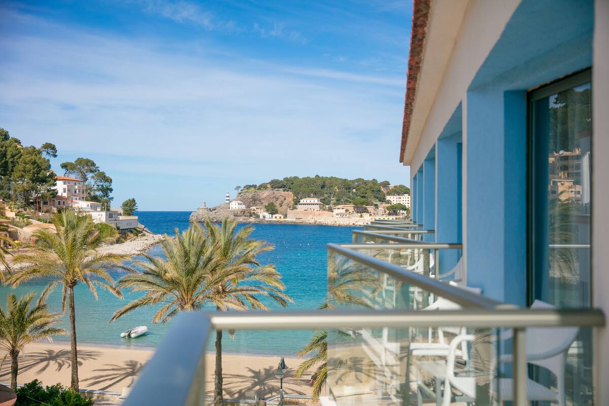 A coastal view from a balcony overlooking a beach with palm trees, a clear blue sea, and buildings in the background.