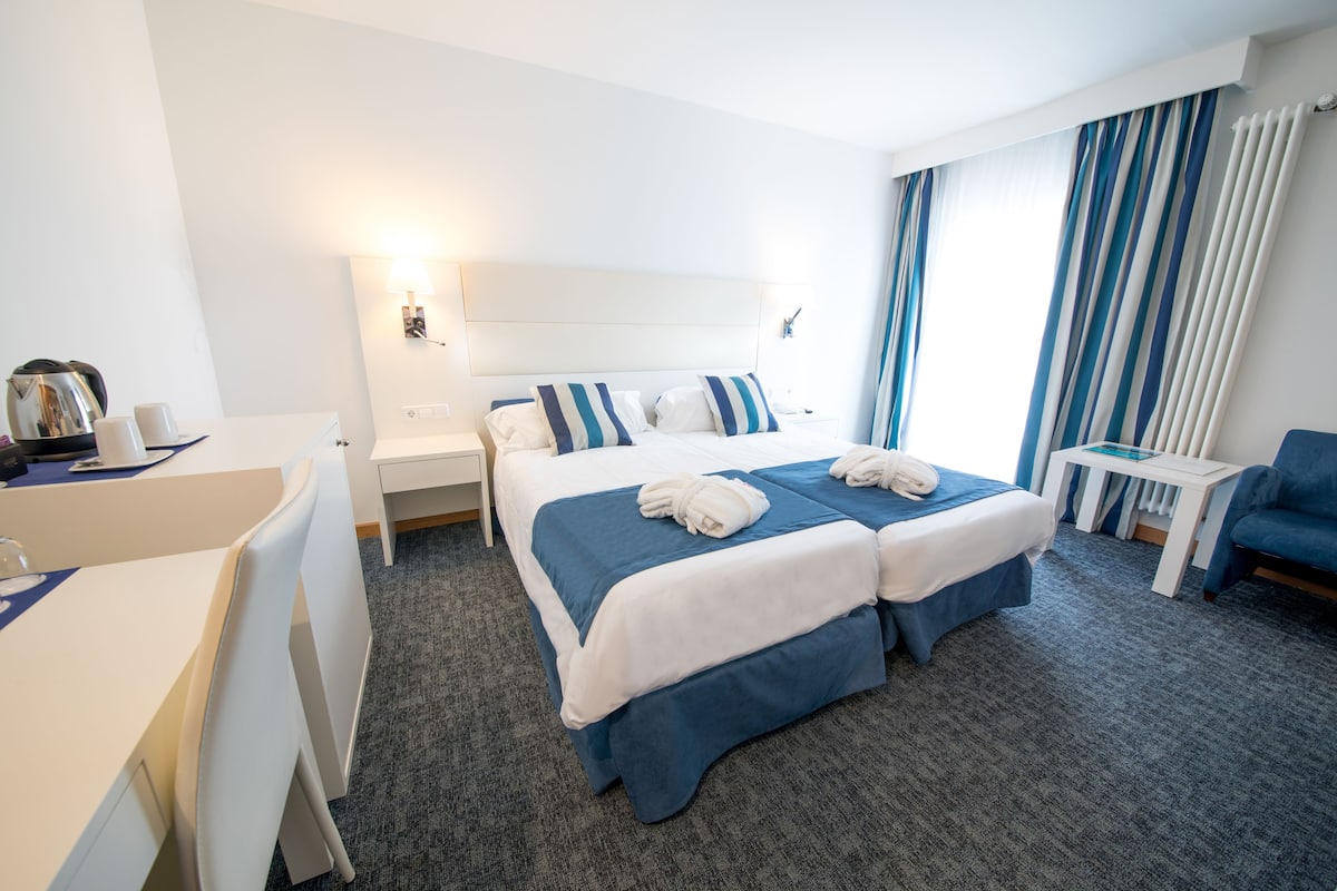 A hotel room with two beds, a desk, a chair, a small table, and a blue armchair.