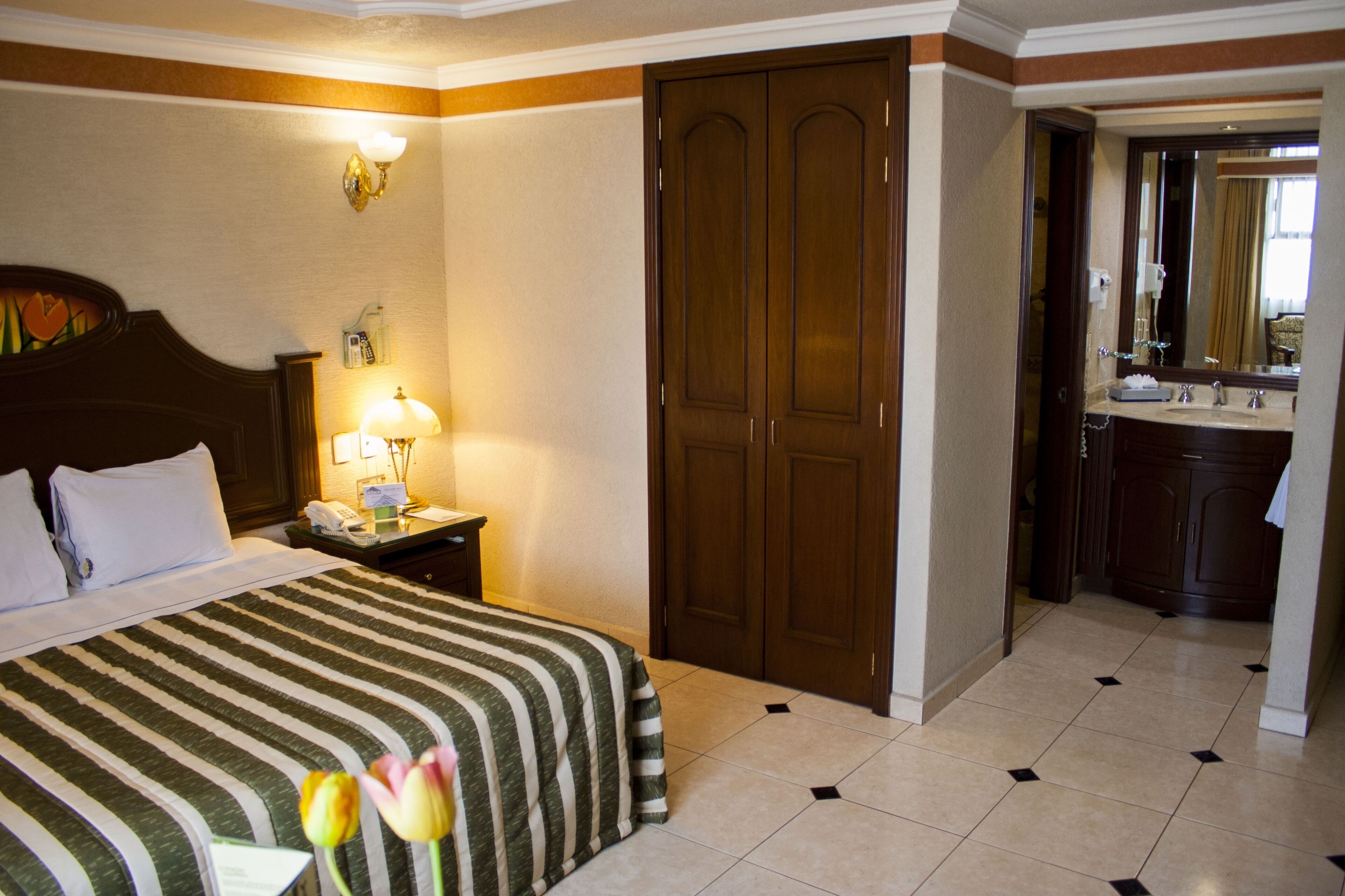 Habitación de hotel con cama, mesita de noche, lámpara, espejo, baño con lavamanos y espejo, y una puerta.