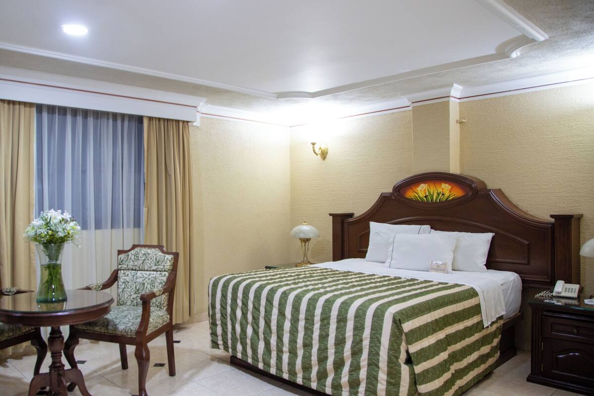 Habitació d'hotel amb un llit, una taula de treball, una cadira, un vas amb flors i una finestra amb cortines.