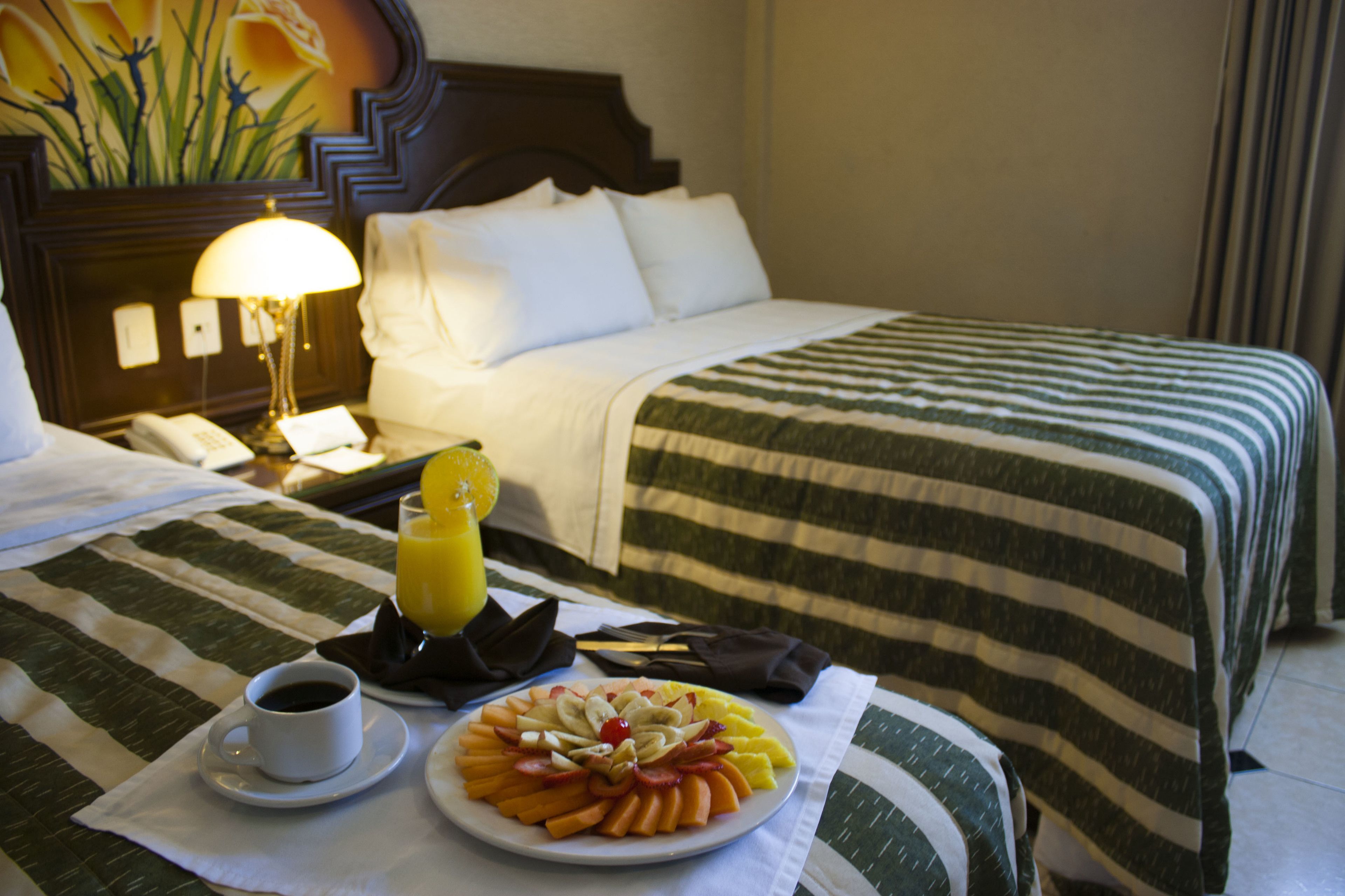 Habitación de hotel con una cama bien hecha, una mesita de noche con lámpara, un plato de fruta, una taza de café y un vaso de zumo de naranja.