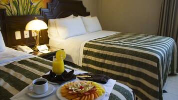 Habitación de hotel con una cama bien hecha, una mesita de noche con lámpara, un plato de fruta, una taza de café y un vaso de zumo de naranja.