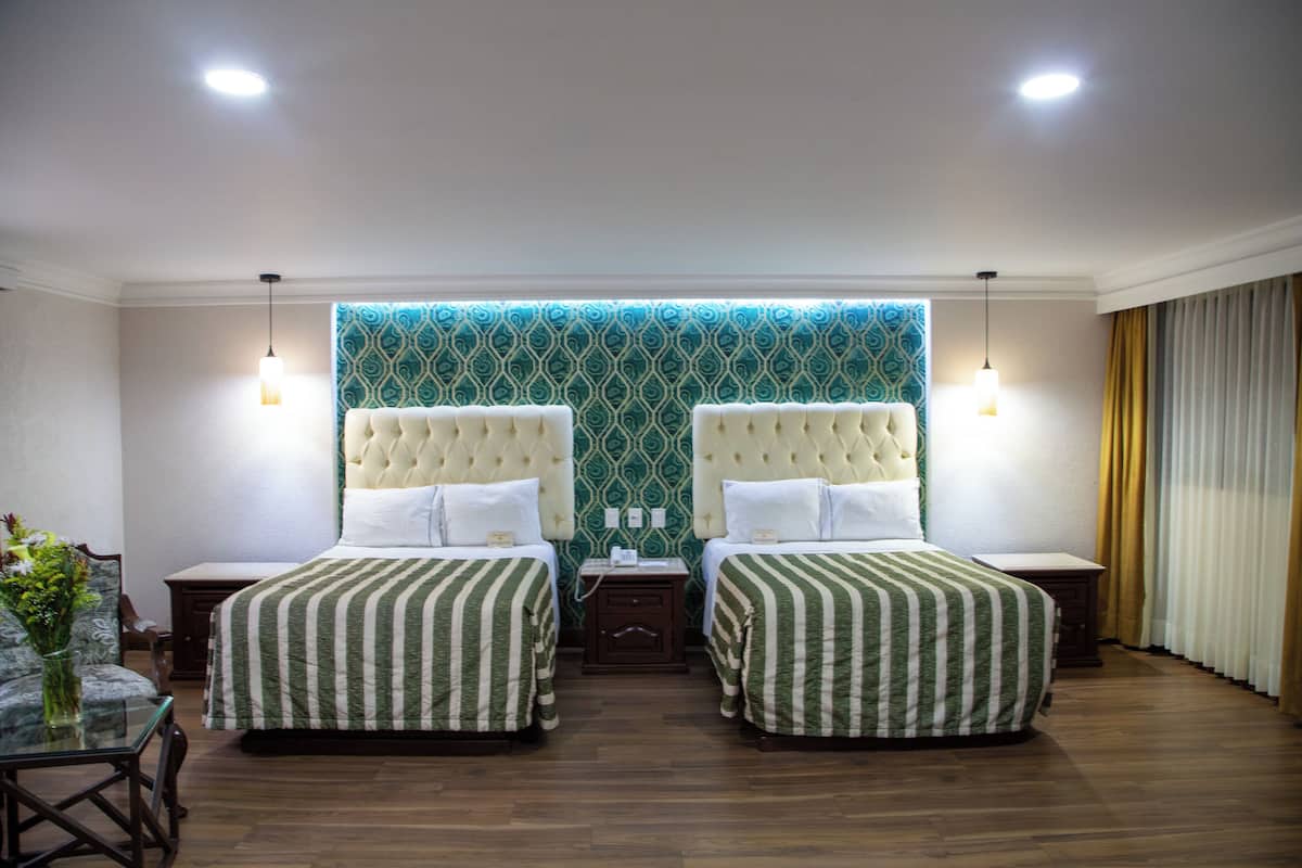 Habitació d'hotel amb dos llits, una tauleta de nit, una taula amb un vas de flors i un mur amb un disseny decoratiu.