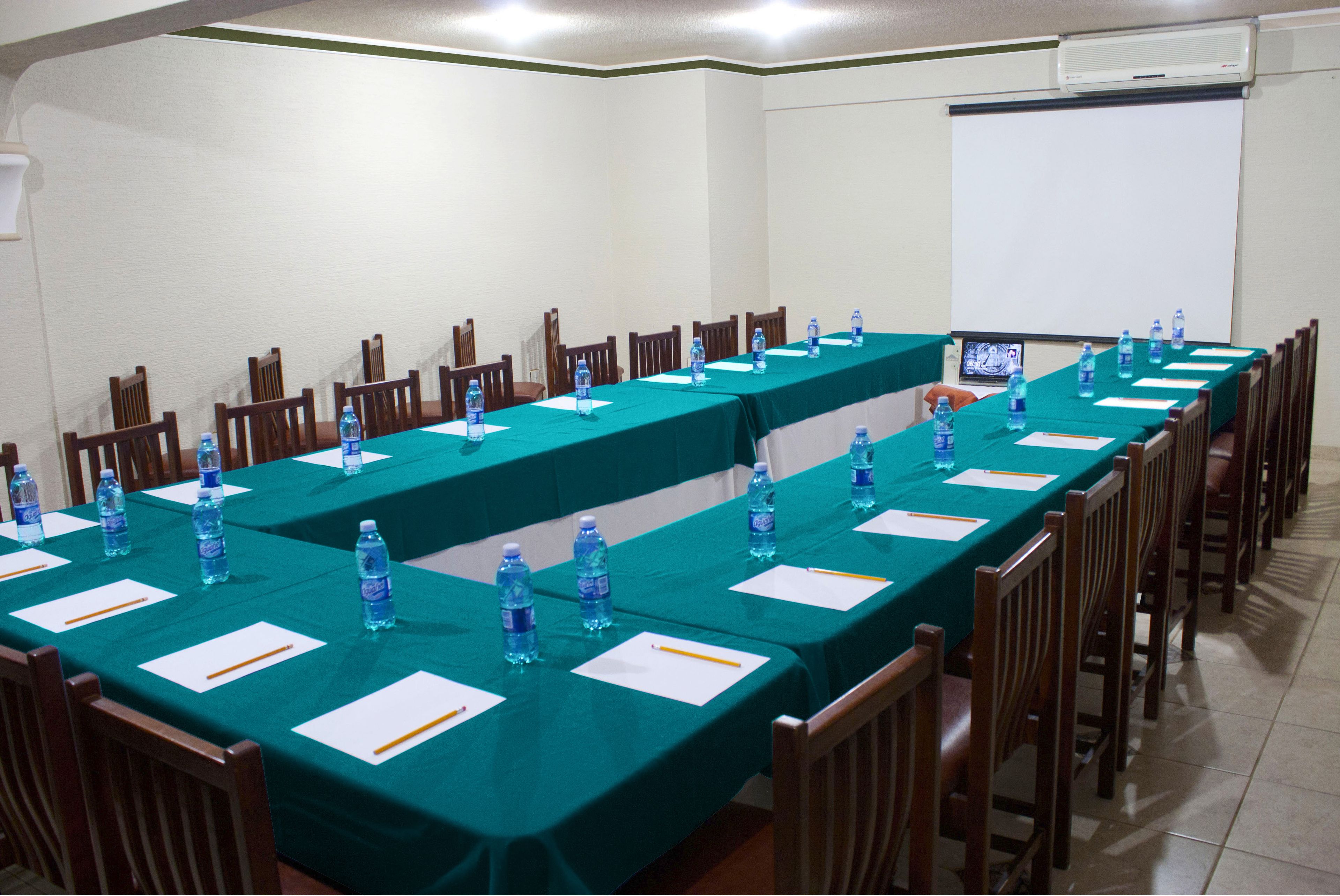 Un salón de conferencias con largas mesas, sillas, botellas de agua y una pantalla para proyector.