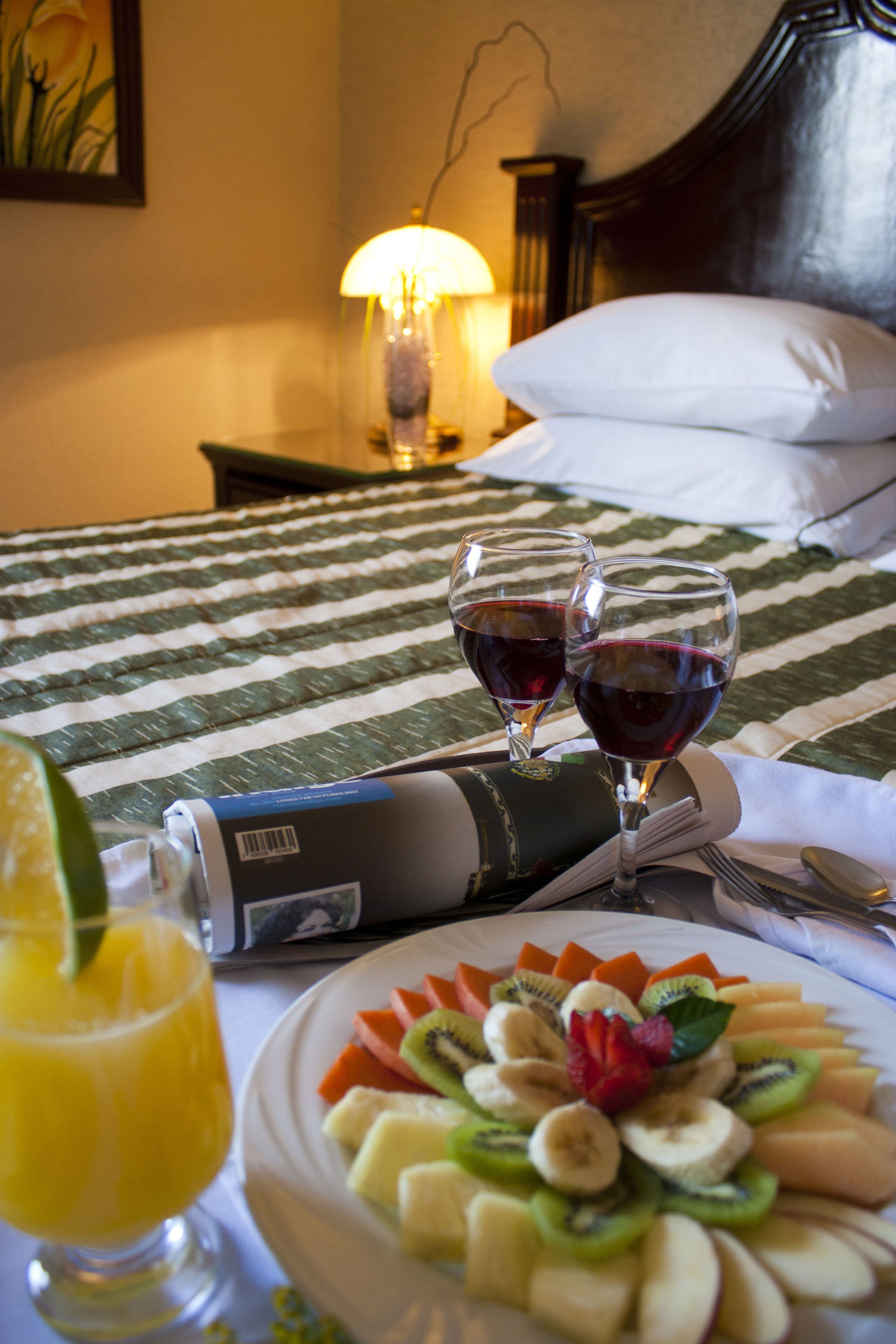 Cama bem arrumada com colcha listrada, abajur e prato com frutas e duas taças de vinho tinto sobre a mesa de cabeceira.