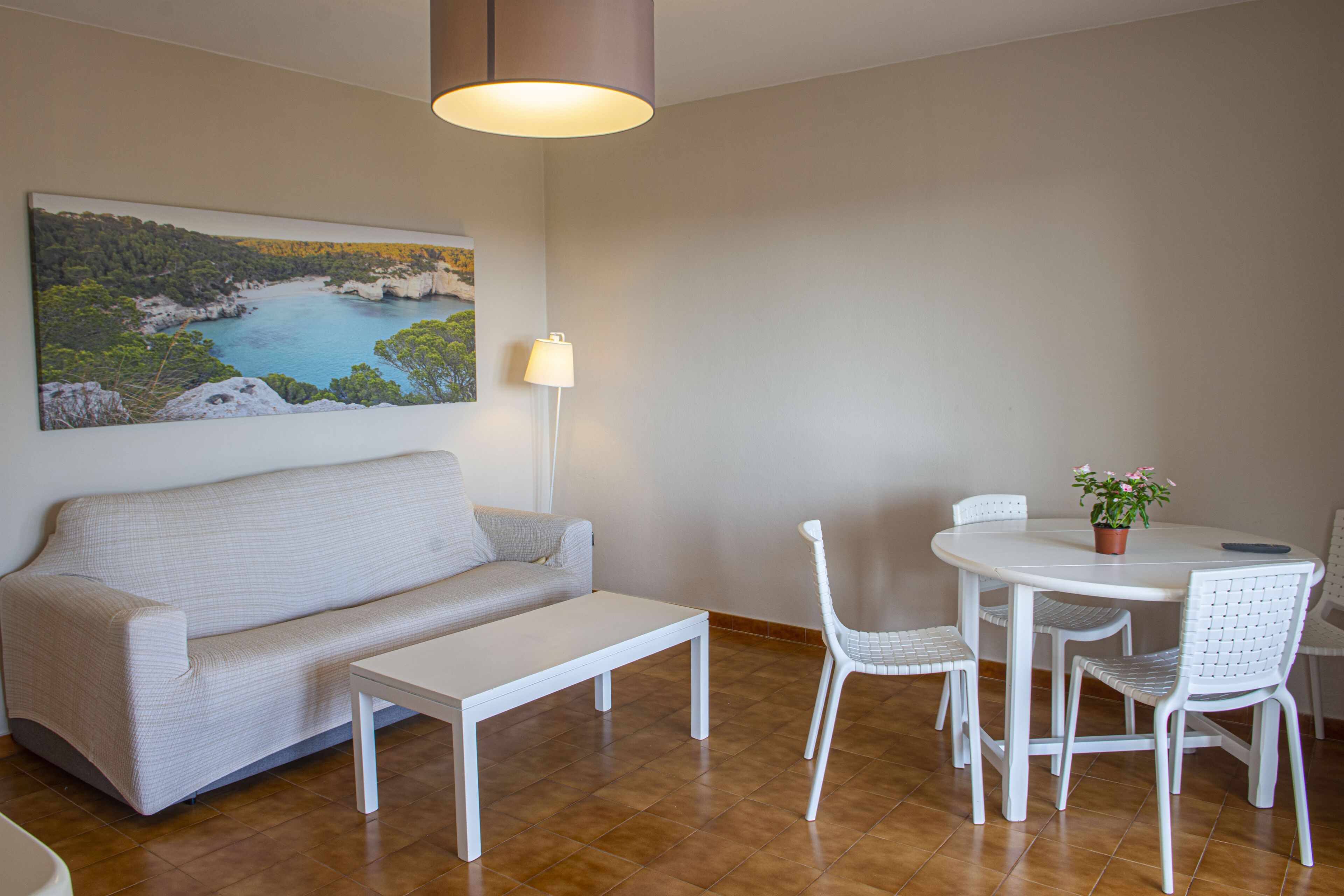 Una sala moderna con un sofá, una mesa de centro, una mesa de comedor con sillas, una lámpara y un cuadro de un paisaje costero.