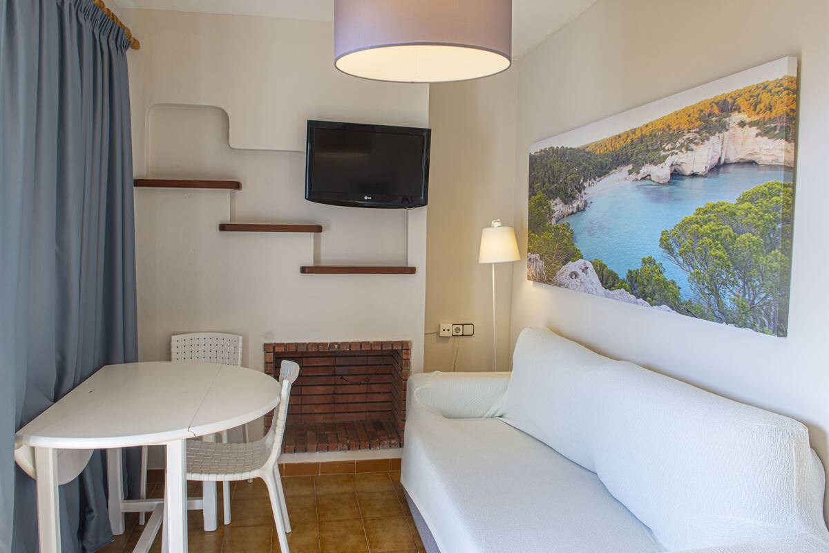 A room with a white table, a white sofa, a TV mounted on the wall, and a painting of a coastal landscape.