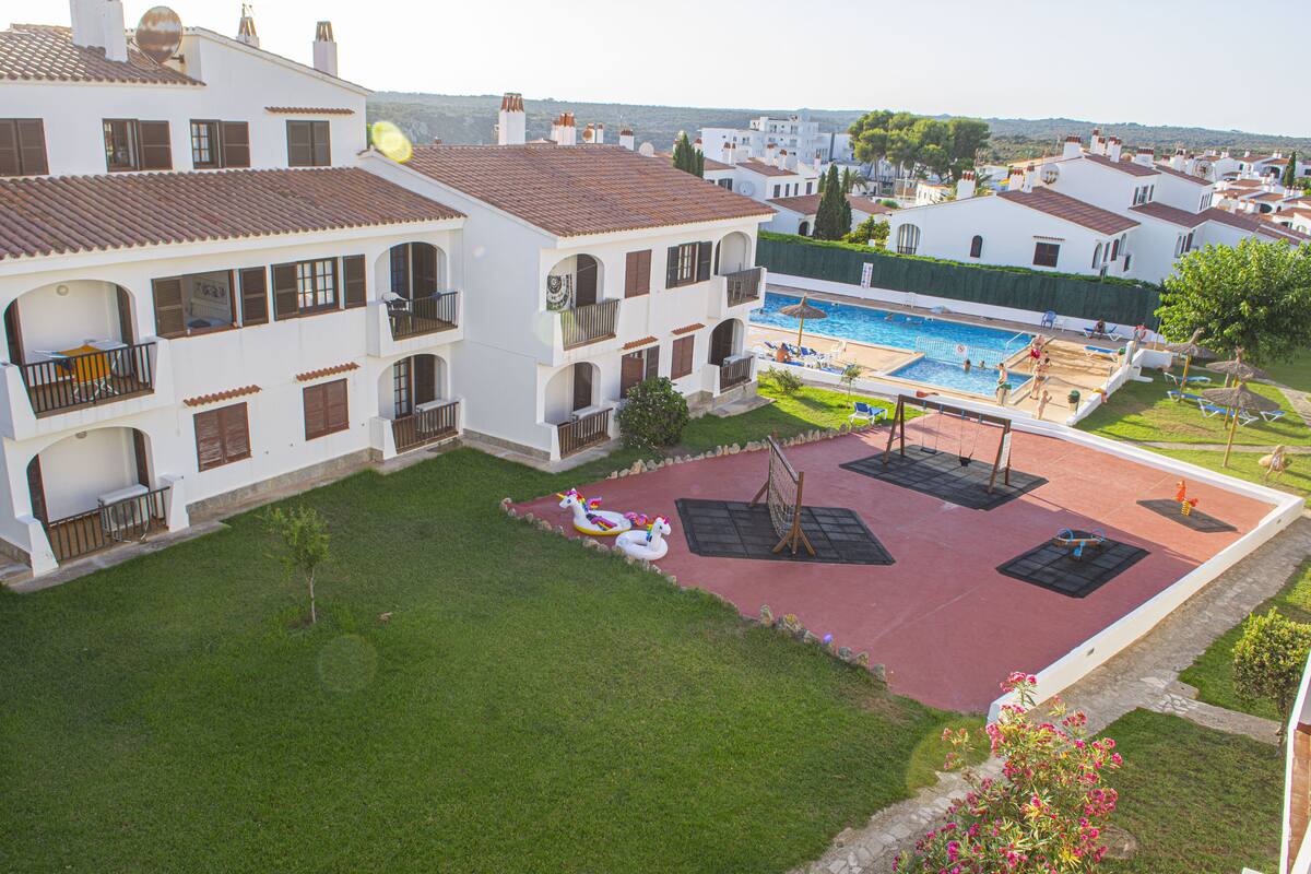 A white building with a swimming pool, a tennis court, and a small garden.