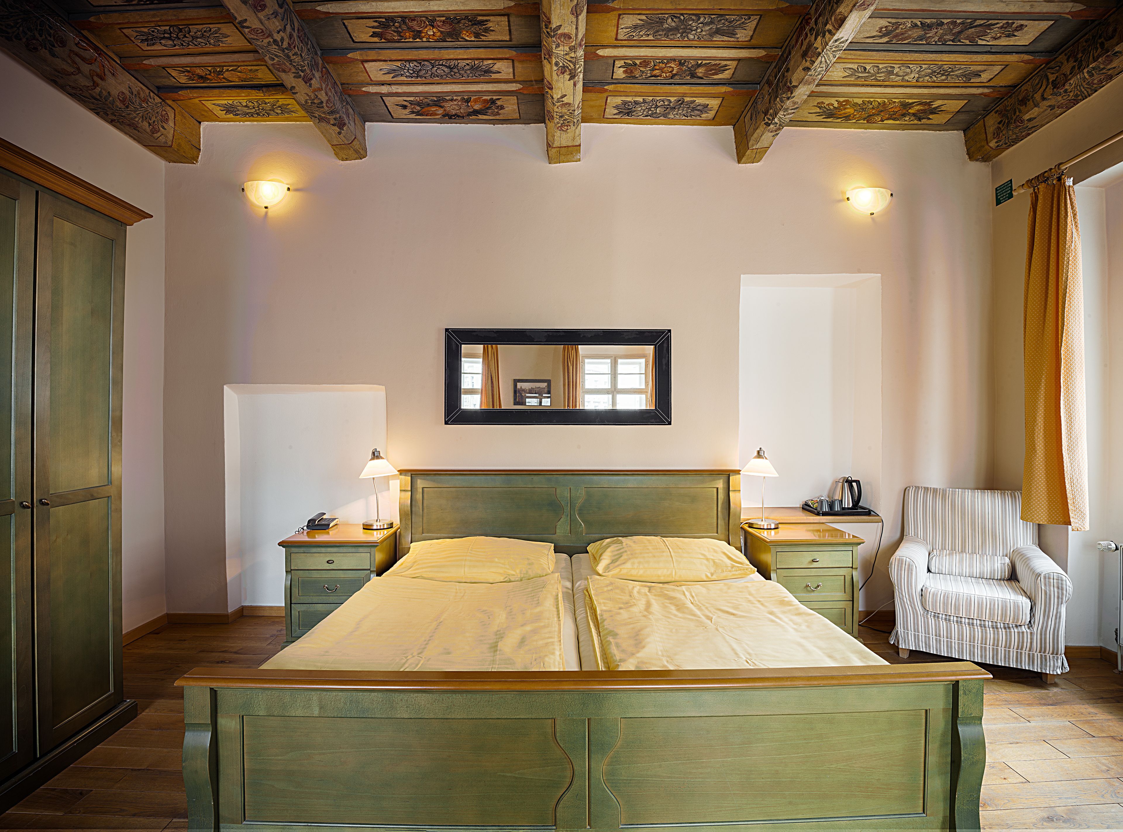 A bedroom with a double bed, two bedside tables, a chair, a mirror, and a ceiling with intricate woodwork.