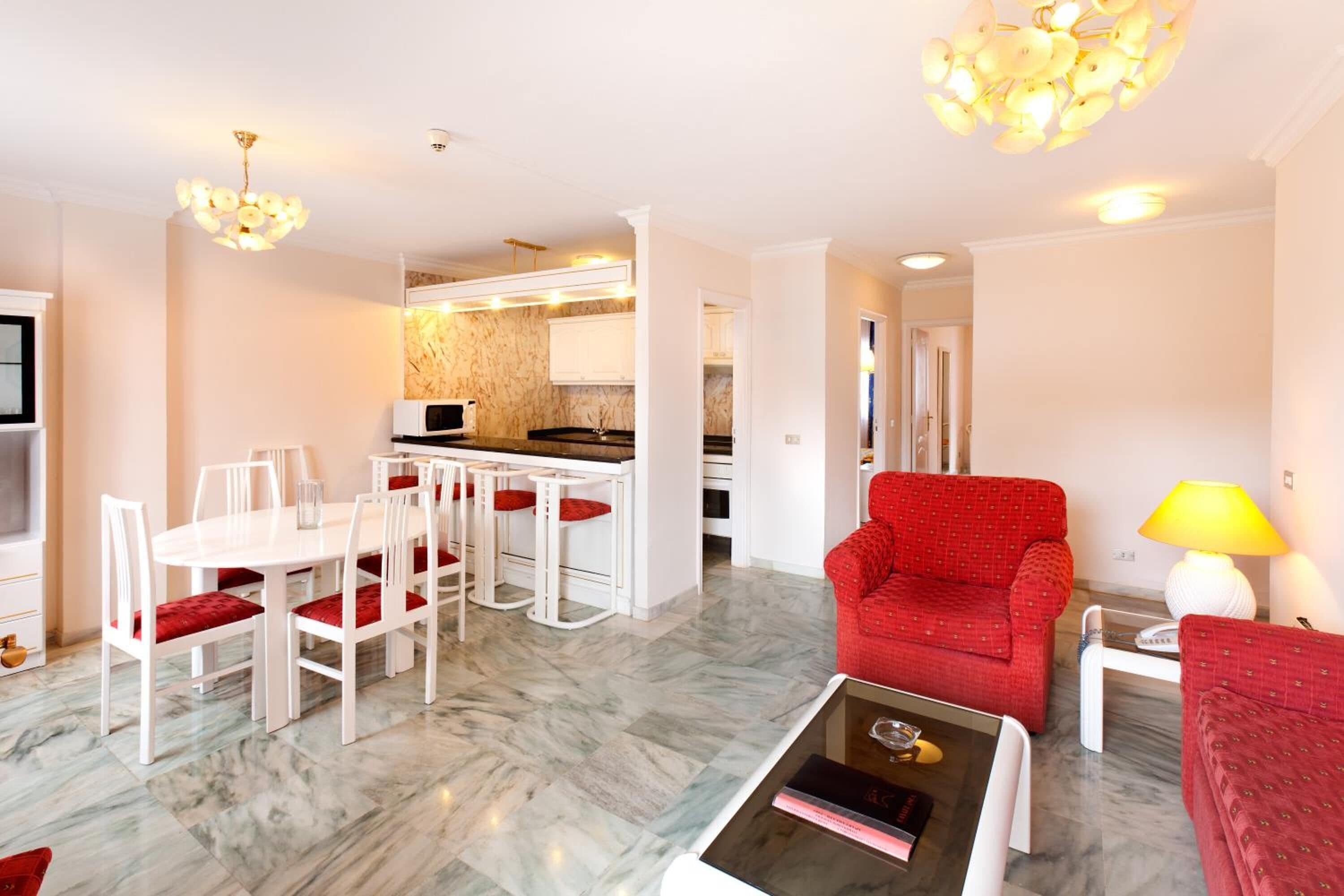 A modern living room with a red armchair, a dining area with white chairs, and a kitchen in the background.