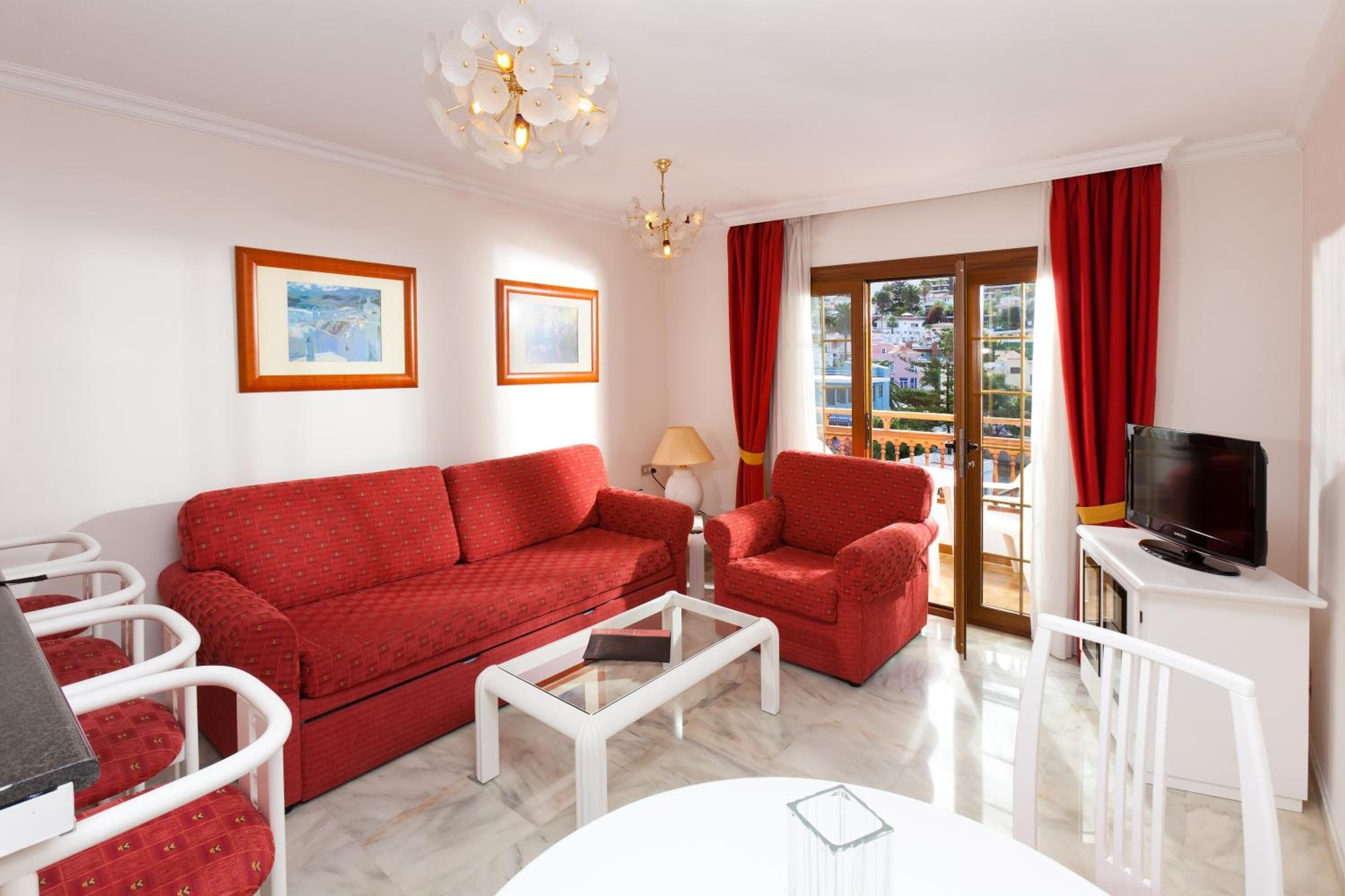 A living room with a red sofa, white armchairs, a coffee table, a TV, and a balcony with red curtains.