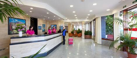 A hotel reception area with a marble counter, reception staff, and potted plants.