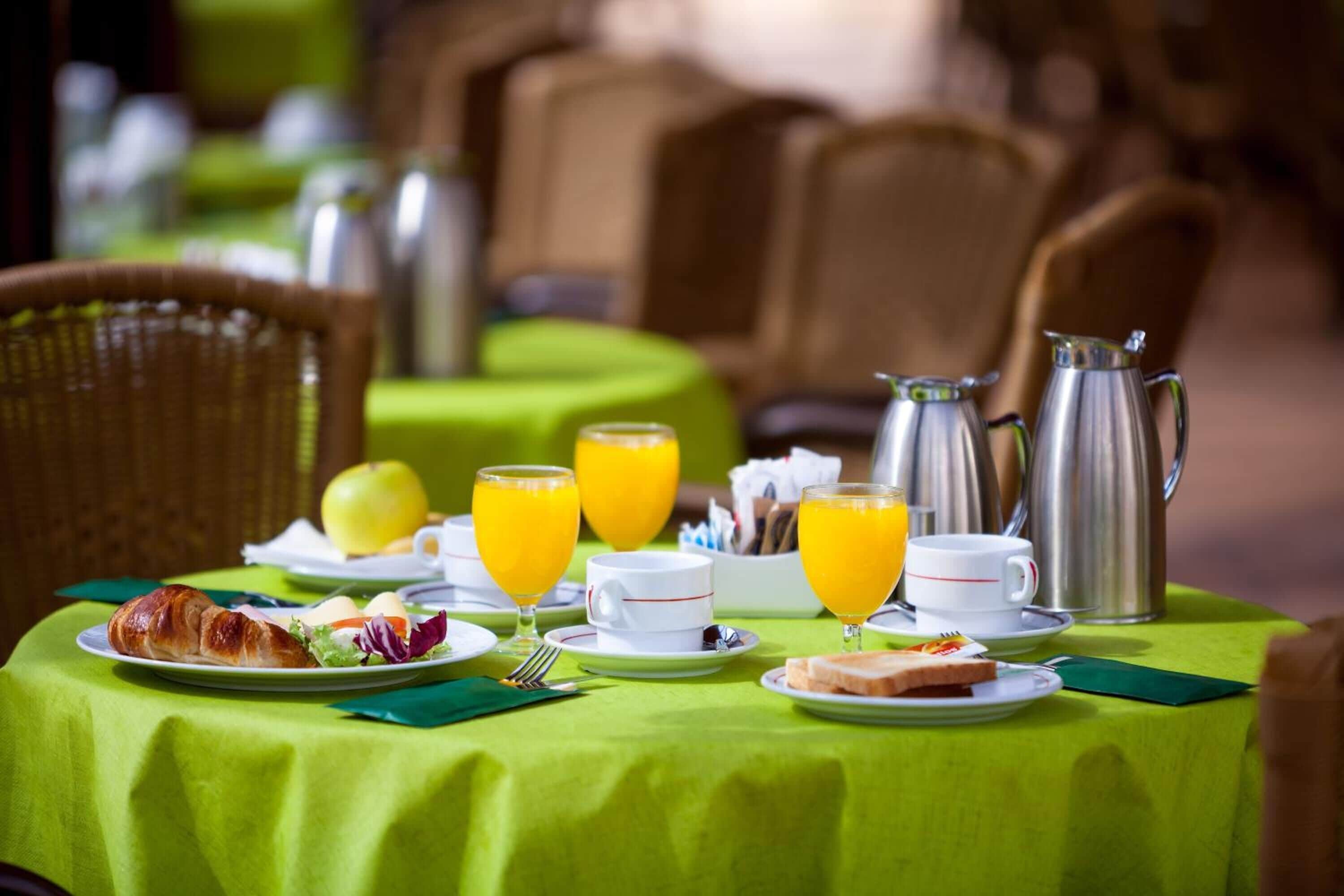 A breakfast table set with croissants, eggs, toast, and orange juice glasses, with a coffee pot and a fruit bowl in the background.