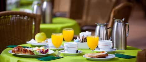 A breakfast table set with croissants, eggs, toast, and orange juice glasses, with a coffee pot and a fruit bowl in the background.