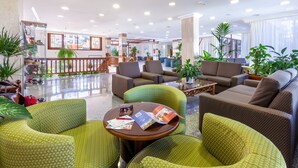 Un vestíbulo amplio con varias zonas de estar, incluyendo sillones tapizados en tonos verde y marrón, una mesa de centro y una mesa de vidrio con revistas y un bolígrafo.