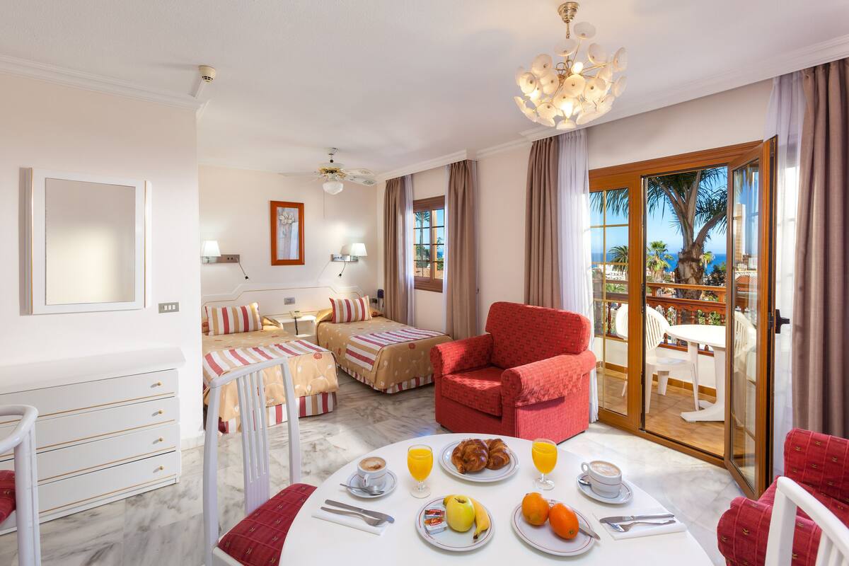 A hotel room with a bed, a red armchair, a dining table with breakfast, and a view of the sea through a glass door.