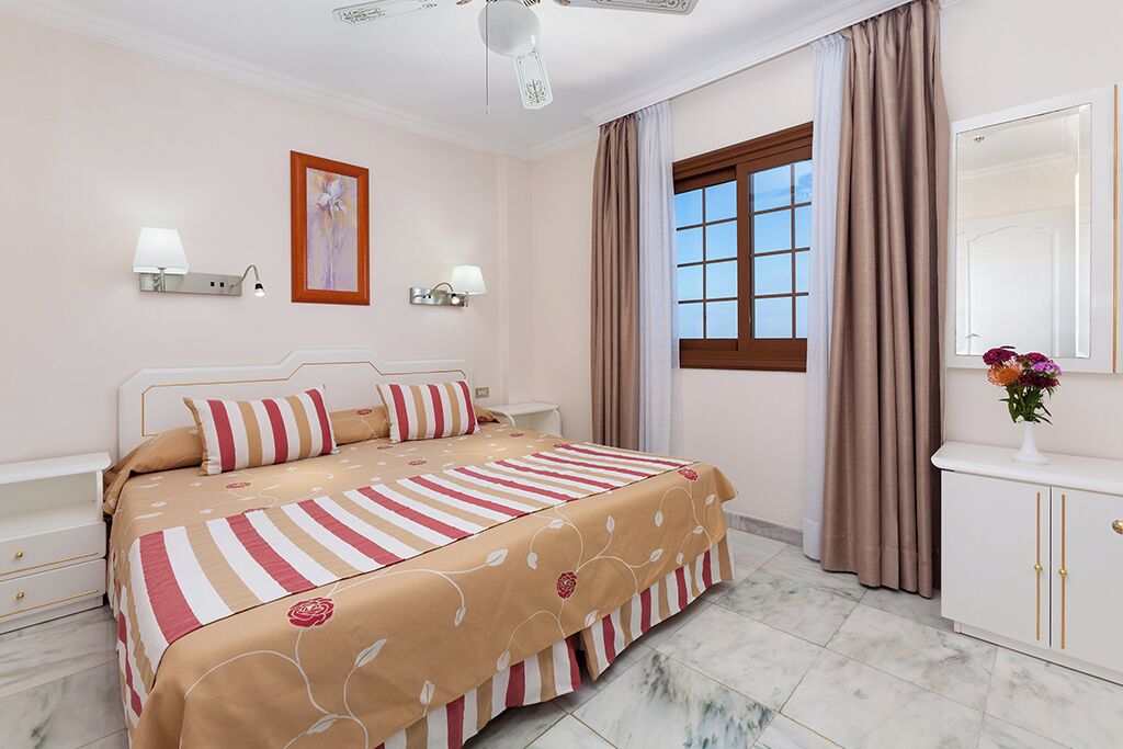 A hotel room with a bed, bedside lamps, a window with curtains, a mirror, and a vase with flowers.