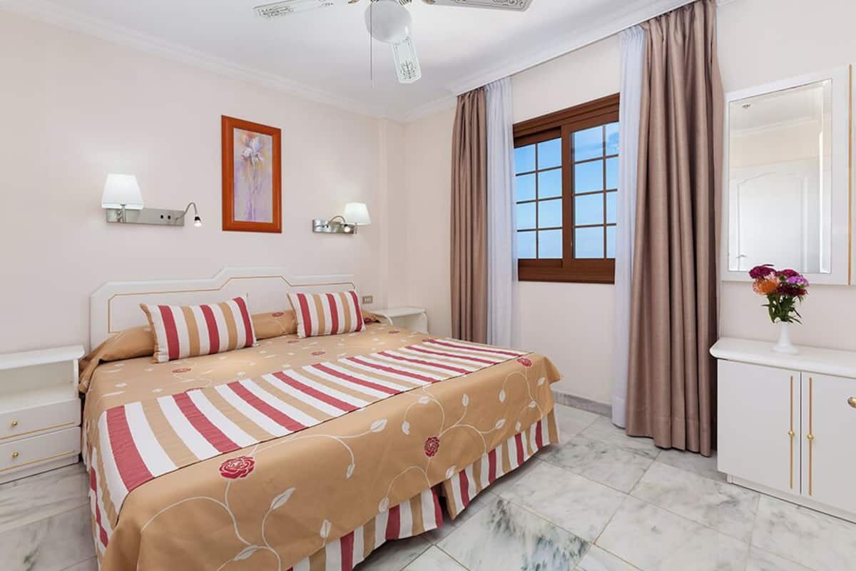 A hotel room with a bed, bedside lamps, a window with curtains, a mirror, and a vase with flowers.
