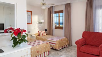 A hotel room with two beds, a red sofa, a ceiling fan, a window with curtains, a desk with a lamp, and a potted plant.