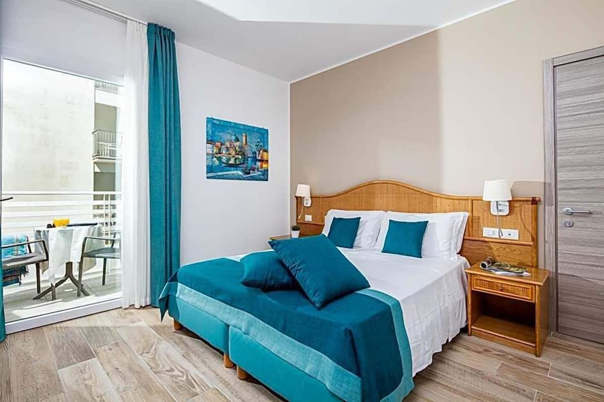 A hotel room with a bed, bedside tables, a window with curtains, a painting on the wall, and a balcony with a table and chairs.