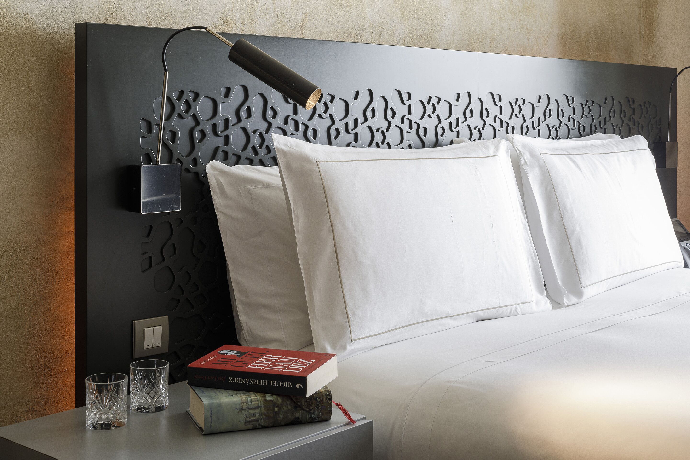Una cama bien hecha con sábanas blancas, un cabecero moderno con diseño geométrico, una lámpara de noche y una pila de libros y gafas sobre la mesita.
