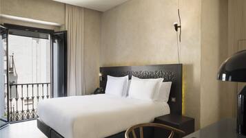 Habitación de hotel con una cama grande, un escritorio y una silla. Cuenta con un balcón con barandilla y vistas a edificios.