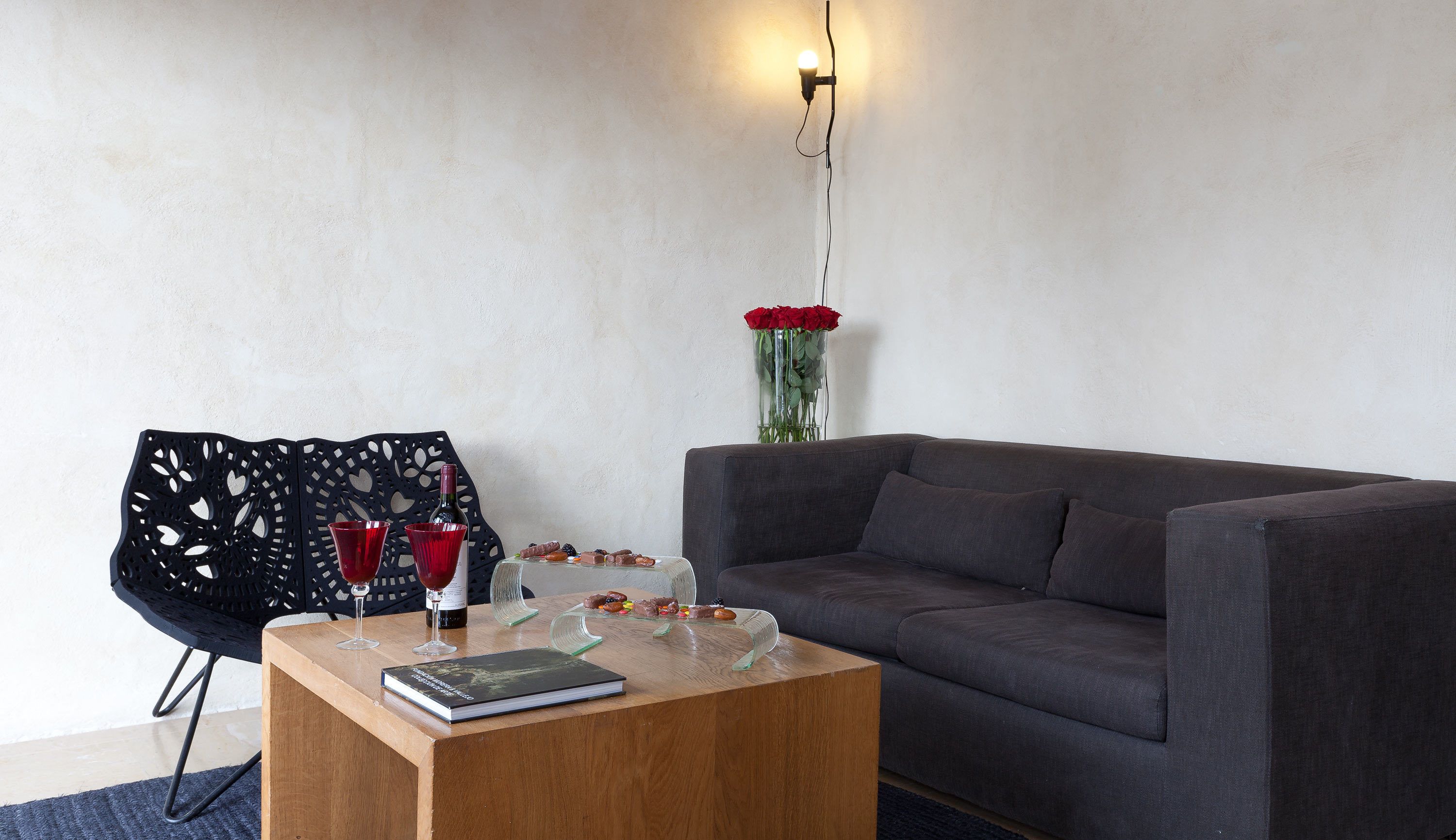 A modern living room with a grey sofa, a wooden coffee table with a book and snacks, a black chair with heart cutouts, and a vase with red roses on the wall.