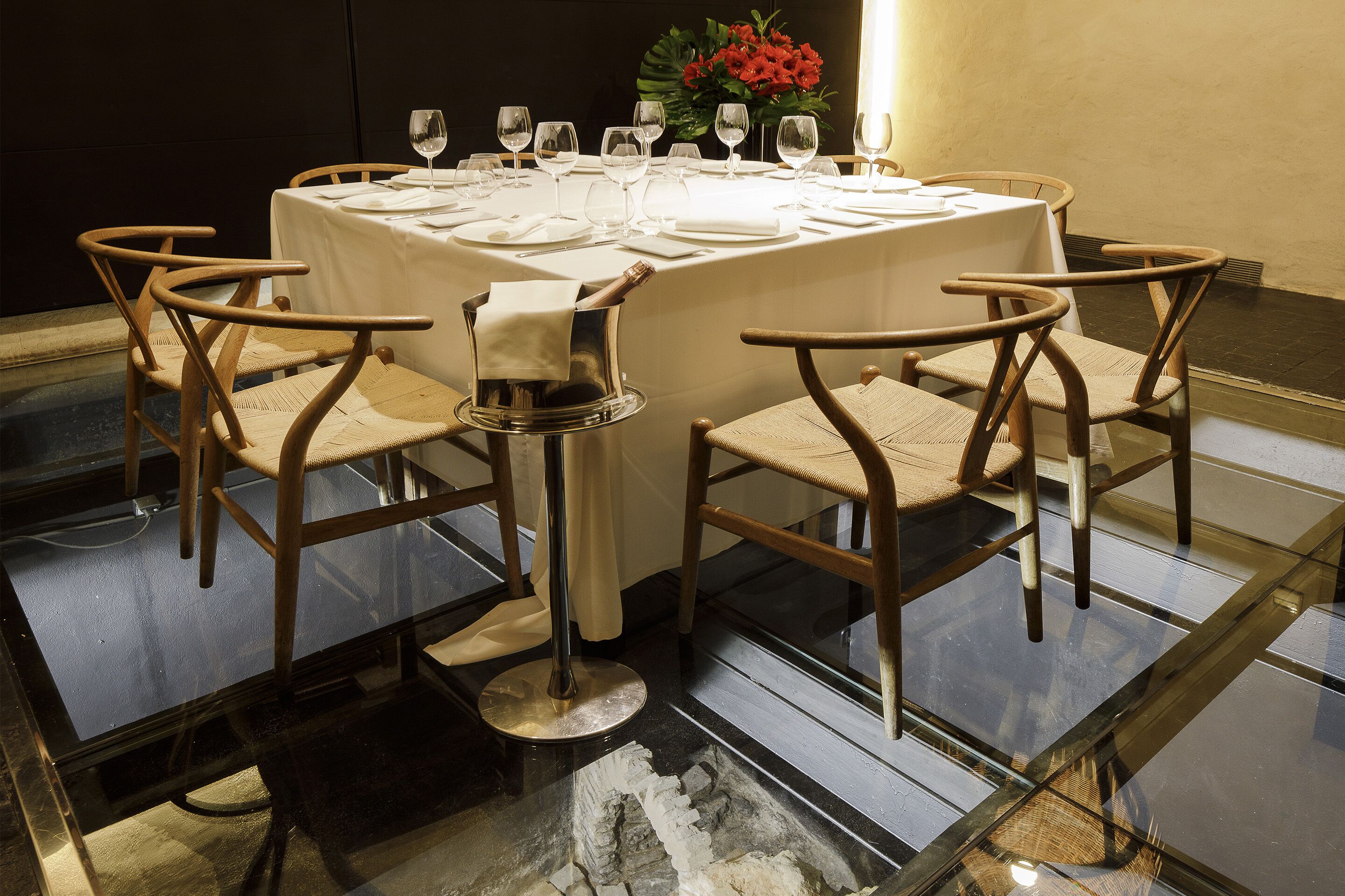 A dining table set for six with white tablecloths, wooden chairs, and wine glasses.