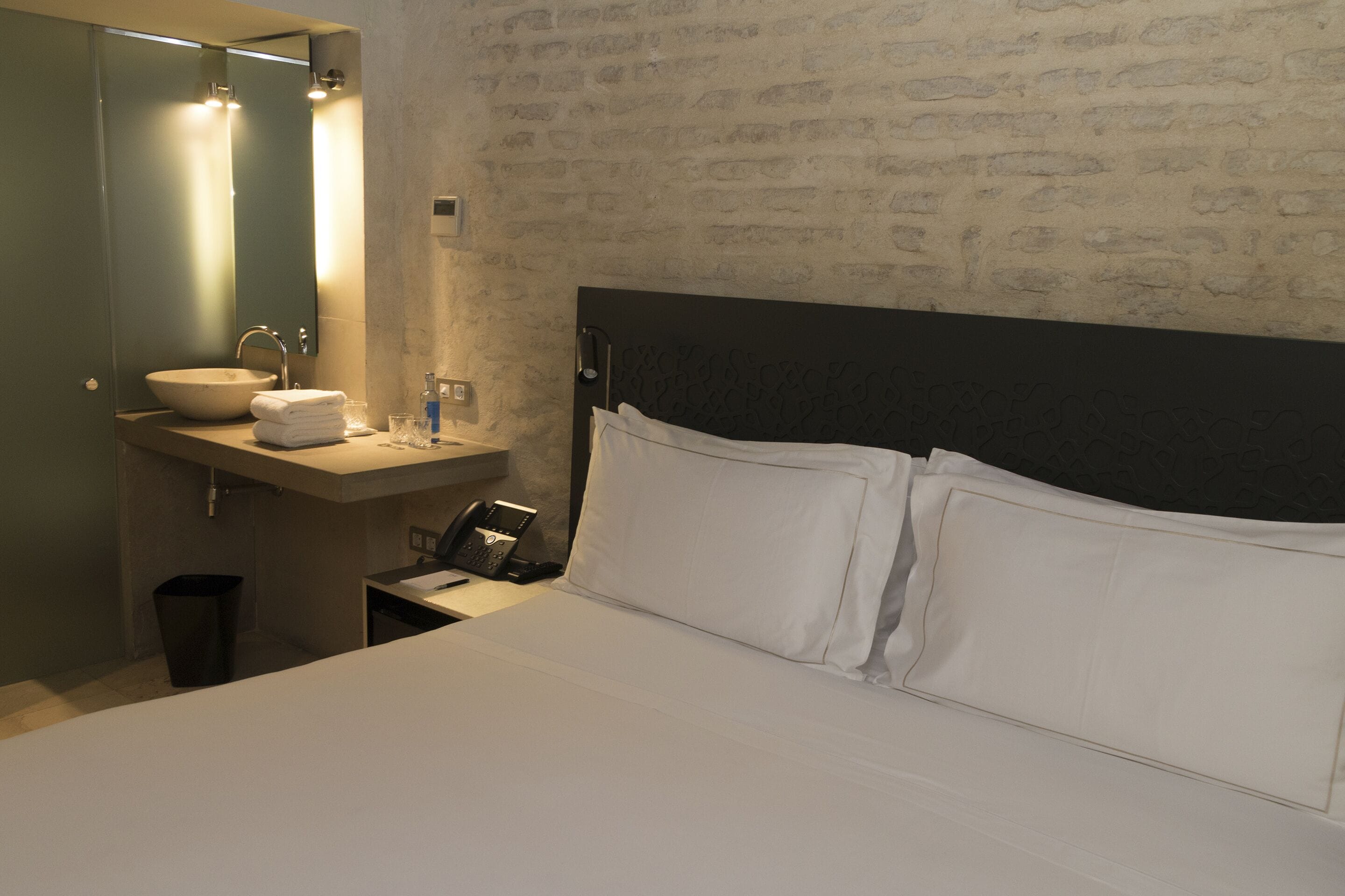 A hotel room with a bed, a bedside table, a nightstand, a sink, and a mirror.