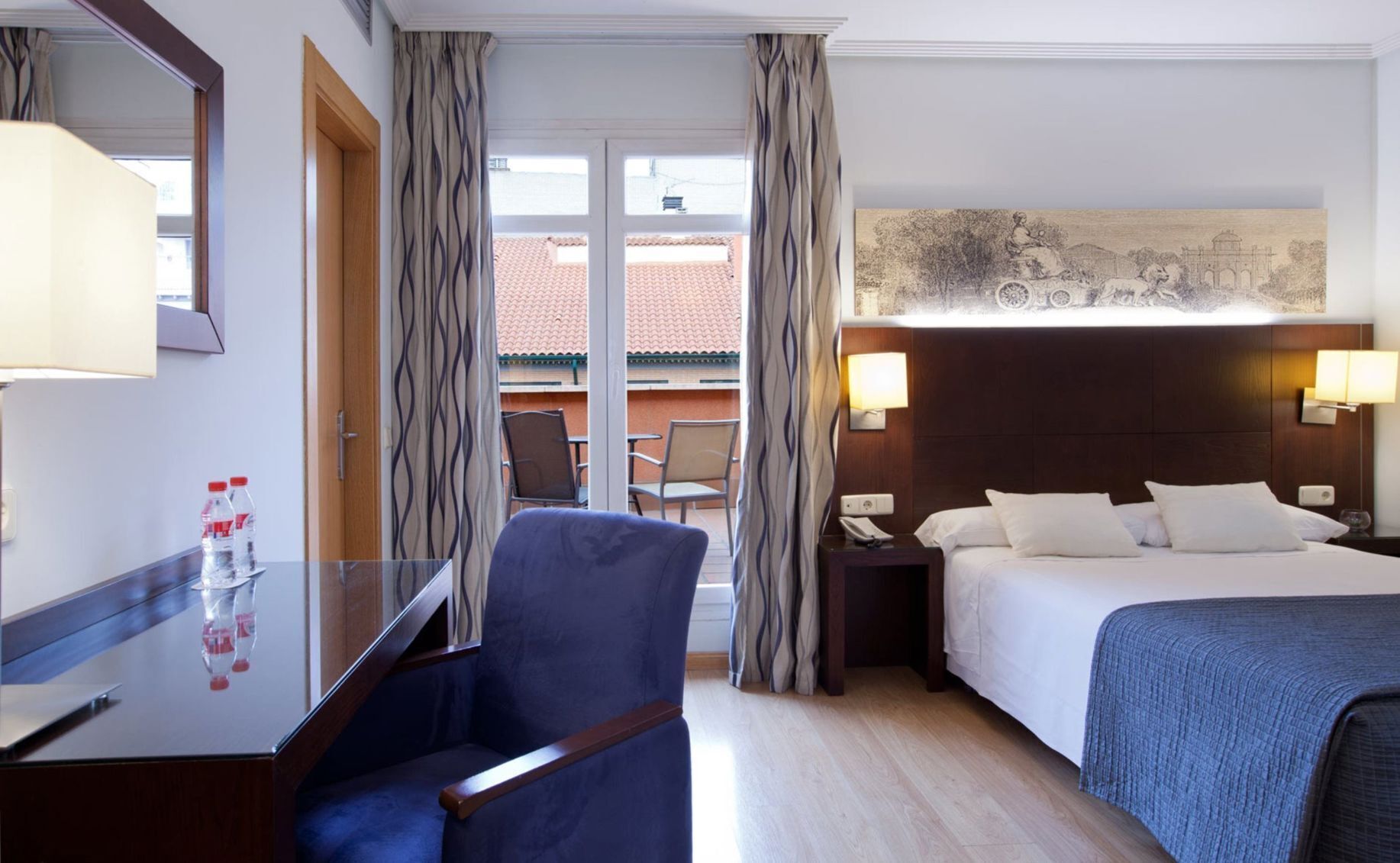 A hotel room with a bed, a desk, a chair, a nightstand, and a view of the outside.