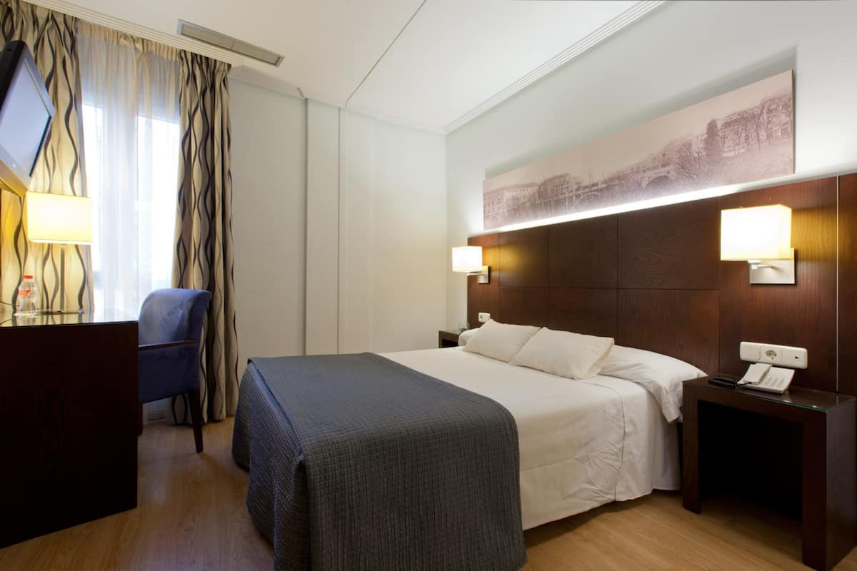 Una habitación de hotel con una cama, lámparas de noche, un escritorio con una silla y un televisor.
