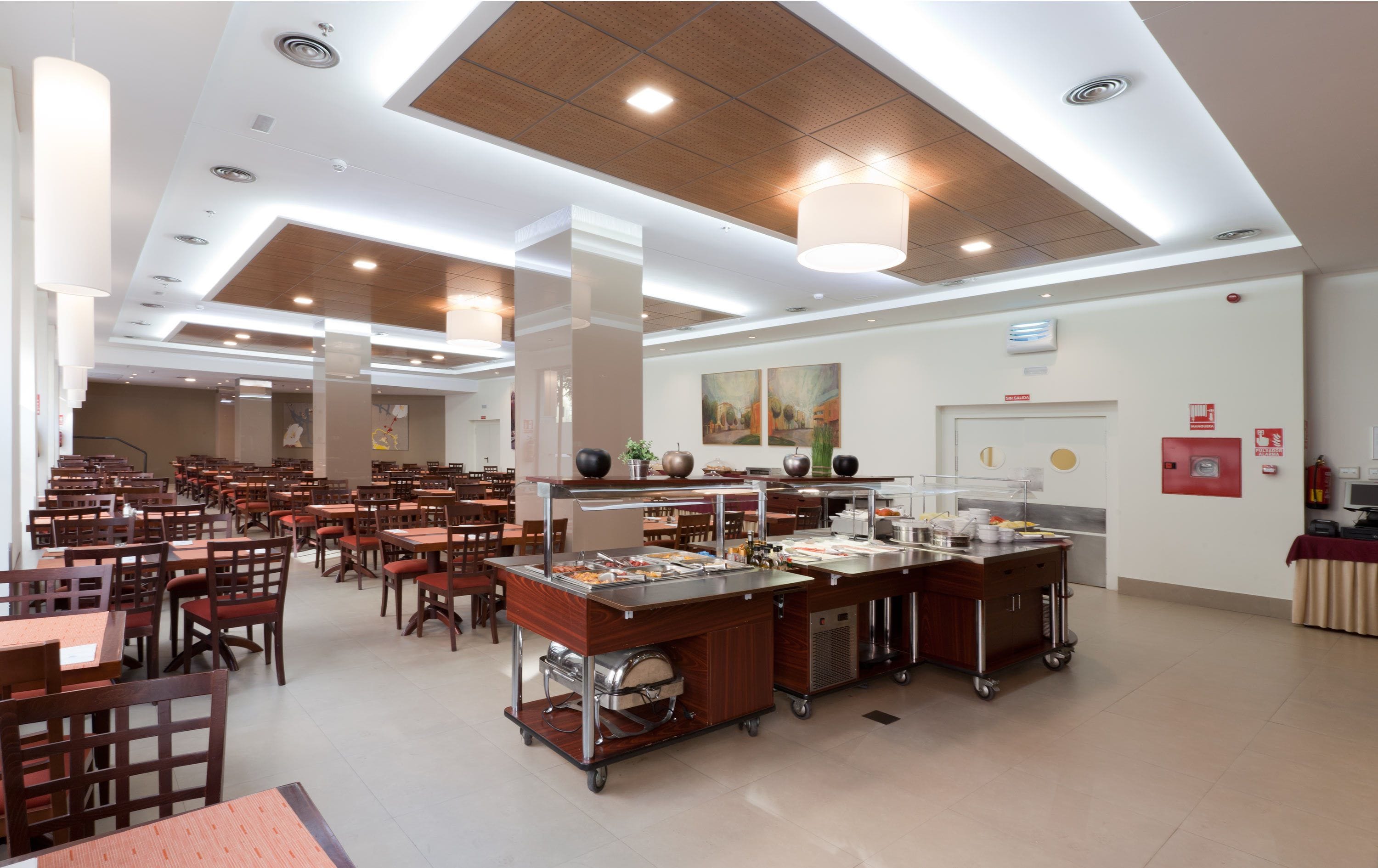 A spacious cafeteria with multiple dining tables and chairs, a buffet area with various food items, and a ceiling with recessed lighting and a wooden panel design.
