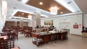 A spacious cafeteria with multiple dining tables and chairs, a buffet area with various food items, and a ceiling with recessed lighting and a wooden panel design.