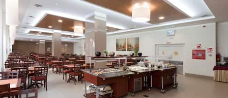 A spacious cafeteria with multiple dining tables and chairs, a buffet area with various food items, and a ceiling with recessed lighting and a wooden panel design.