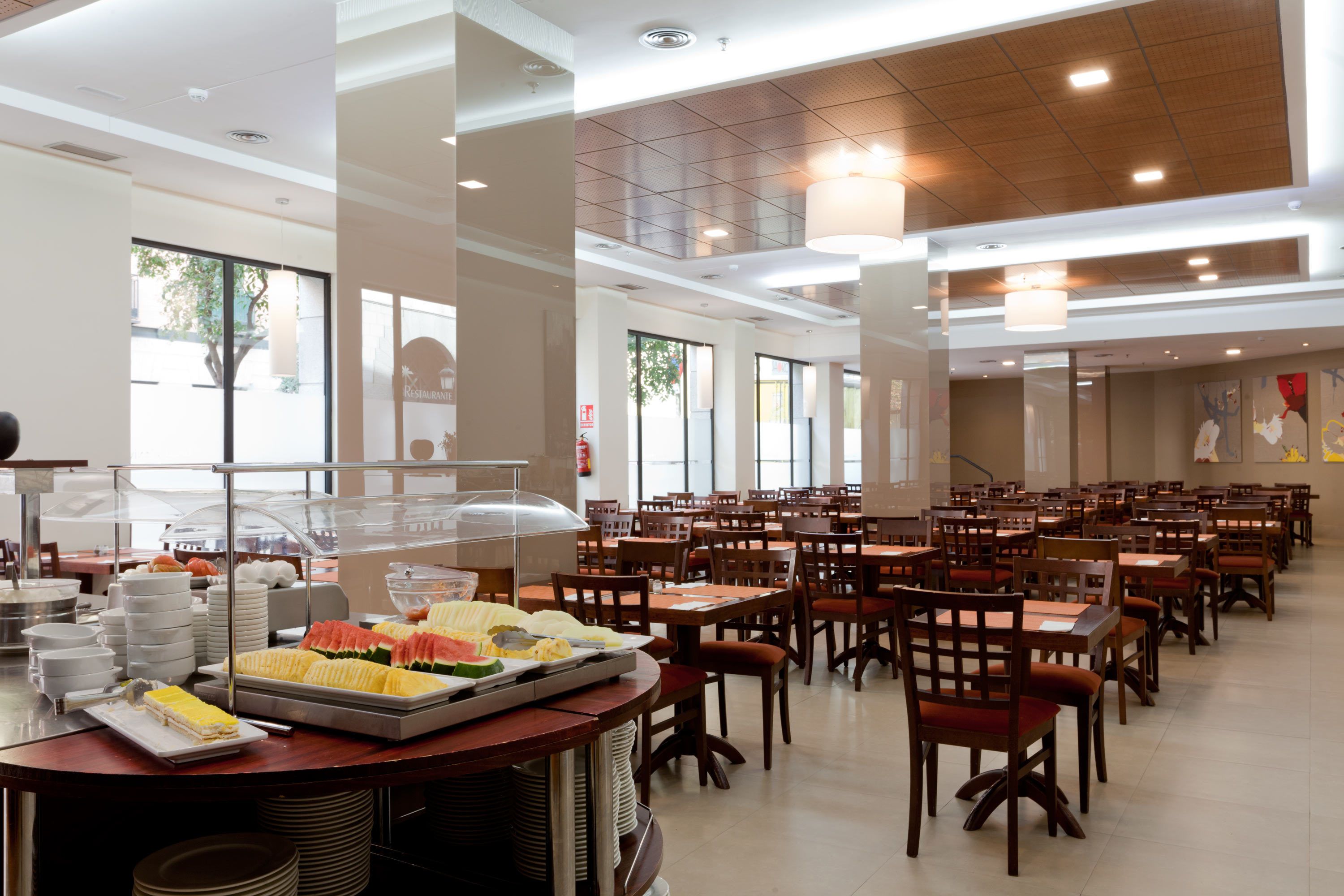 A spacious cafeteria with multiple tables and chairs, a buffet area with various food items, and large windows providing natural light.