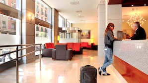 A hotel lobby with a reception desk, red seating, a black suitcase, and a large window overlooking a building. - Hotel Porcel Ganivet (Madrid)